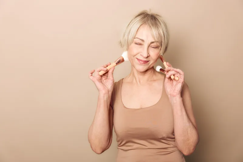 Happy mature woman with short blonde hair holding makeup brushes near face smiling with closed eyes on beige background. Concept for beauty, skincare, cosmetics, and self-care routine.