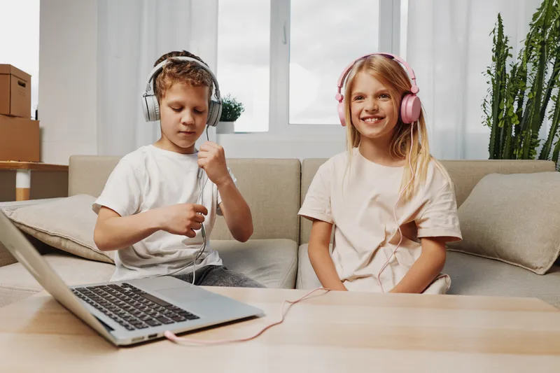 Two happy children with headphones enjoy music and technology at home sitting on a beige sofa near a wooden table with an open laptop. Concept of childhood, entertainment, and modern lifestyle.