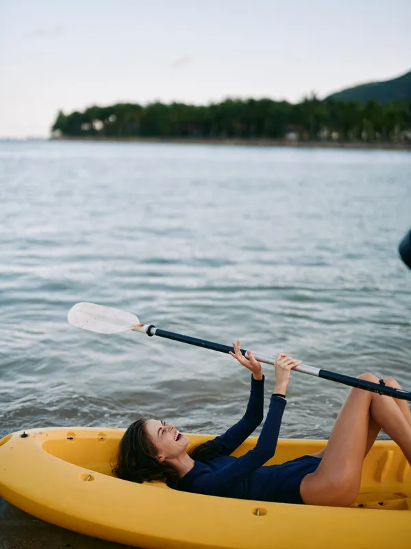 A young woman lies in a yellow kayak near a tranquil shore, raising a paddle overhead with a smile as calm water and trees frame the scene.