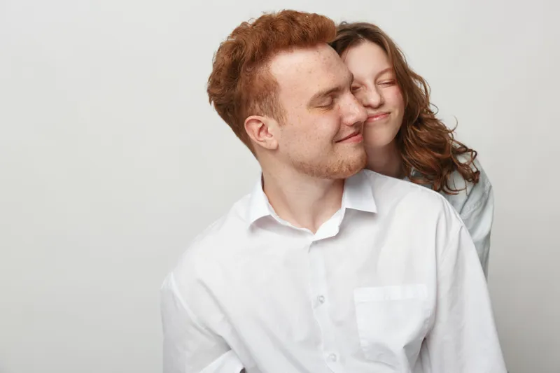 Happy young couple in white shirts embraces with closed eyes and gentle smiles on light background. Concept of love, affection, intimacy, and close relationship between partners.