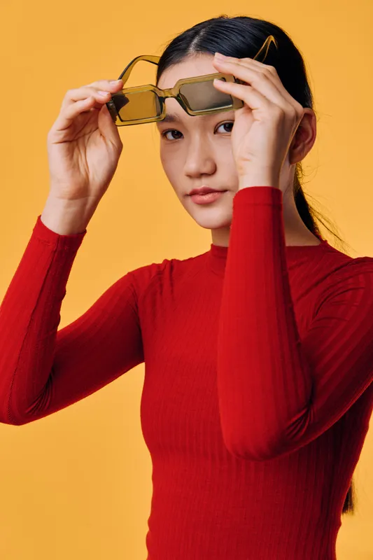 Young woman in red sweater holding sunglasses in front of yellow background. Asian female model with long dark hair posing for fashion portrait with casual expression and minimal makeup.
