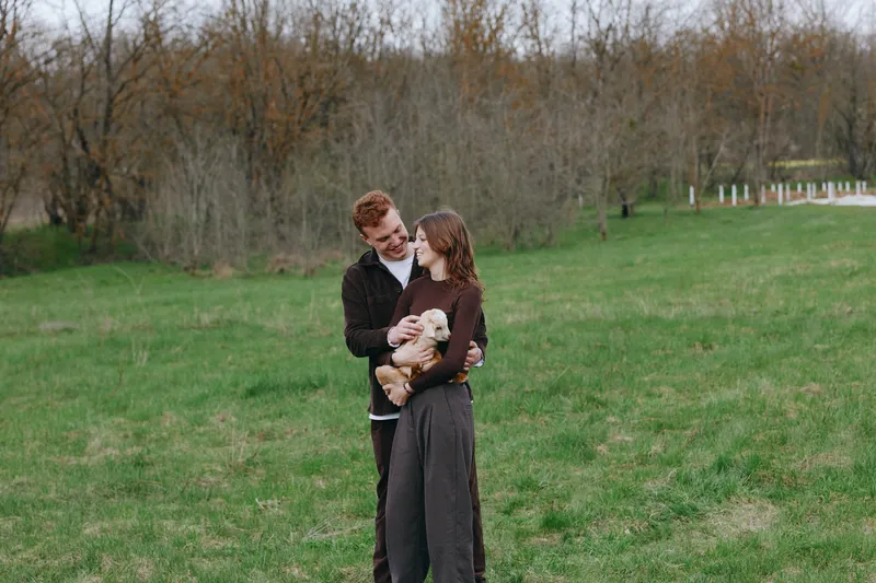 Bright outdoor moment with a couple embracing a goat kid, conveying warm affection and happiness in a sunlit meadow with natural colors and a gentle breeze in a peaceful countryside setting