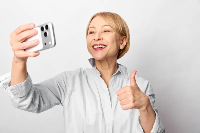 Smiling older woman takes a selfie with a smartphone, giving a thumbs up. Positive mood, technology, and social media lifestyle concept in bright studio today.