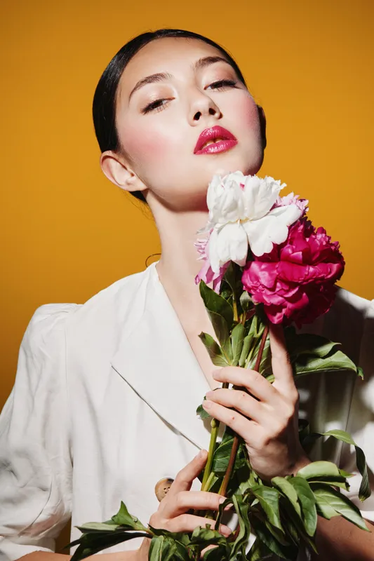 Young woman with slicked hair and red lips hold pink and white peonies in white elegant blazer on warm orange background. Creative beauty and fashion concept.