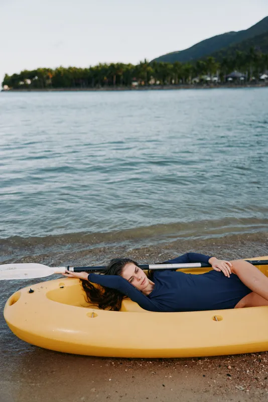 A person lies on a bright yellow raft by calm shoreline, sunlit water, relaxed pose and casual clothing, creating a tranquil outdoor scene of leisure and reflection.