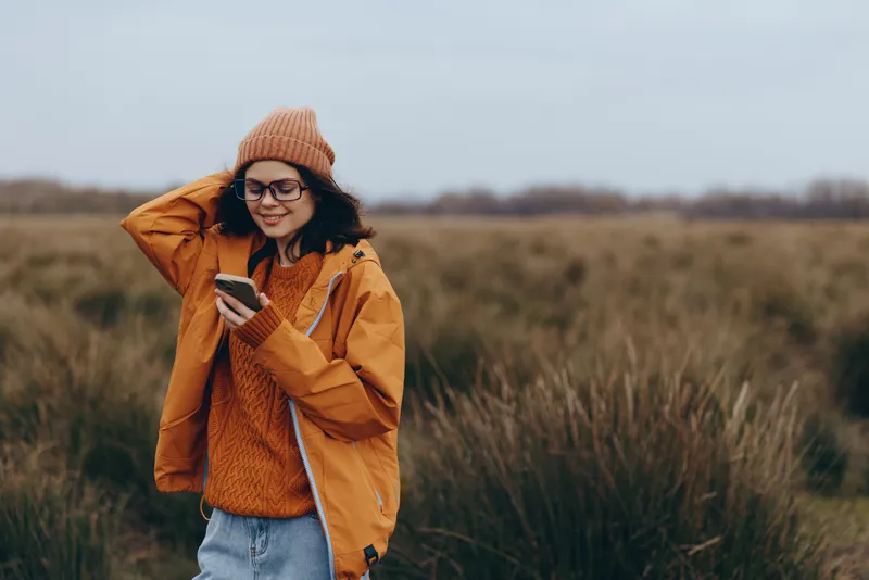 Woman outdoors in an autumn field, wearing orange jacket and knit hat, smiling and looking at a phone, lifestyle moment in warm soft natural light, casual travel mood today