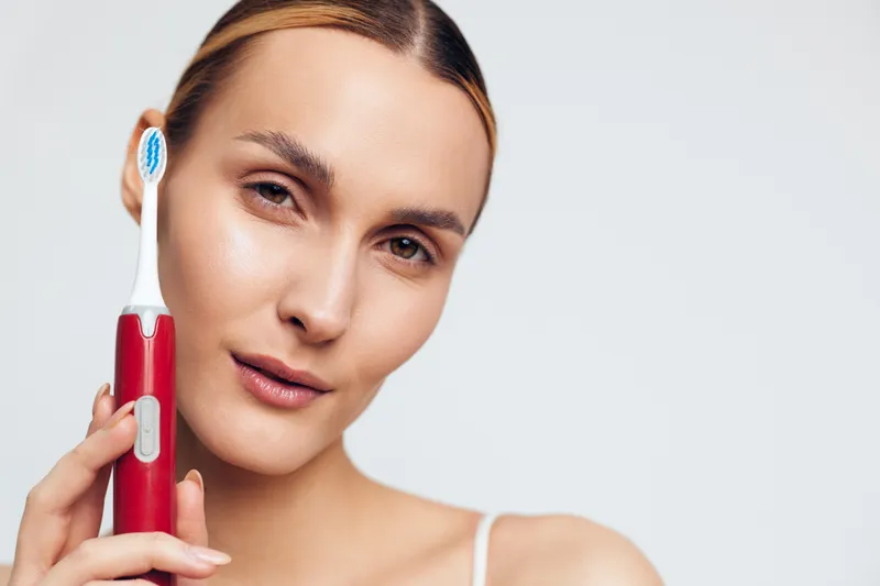 Young woman with brunette hair holds a red electric toothbrush near her face while looking confidently at the camera against a plain light background.
