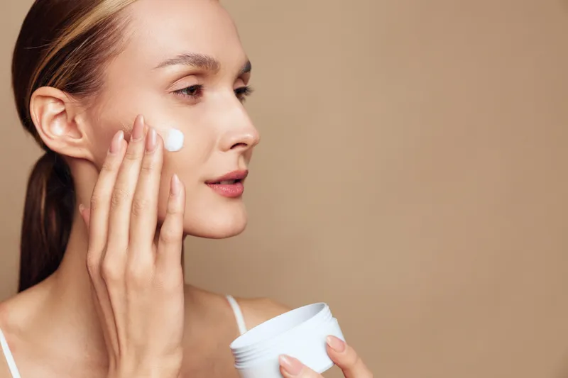 Young woman applying moisturizing cream on face, skincare routine, smooth skin, natural makeup, beige background, close-up, beauty concept, self-care, wellness, gentle touch