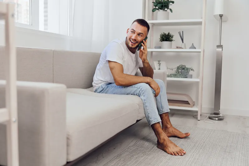 Smiling man sitting on a couch, engaged in a phone call, surrounded by a bright and modern living space, showcasing comfort and connection.