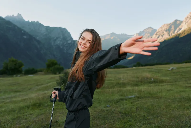 A cheerful hiker stands with arms outstretched in a scenic valley beneath rugged mountains. Bright daylight and an expansive sky create a sense of freedom, adventure, and positive energy in this