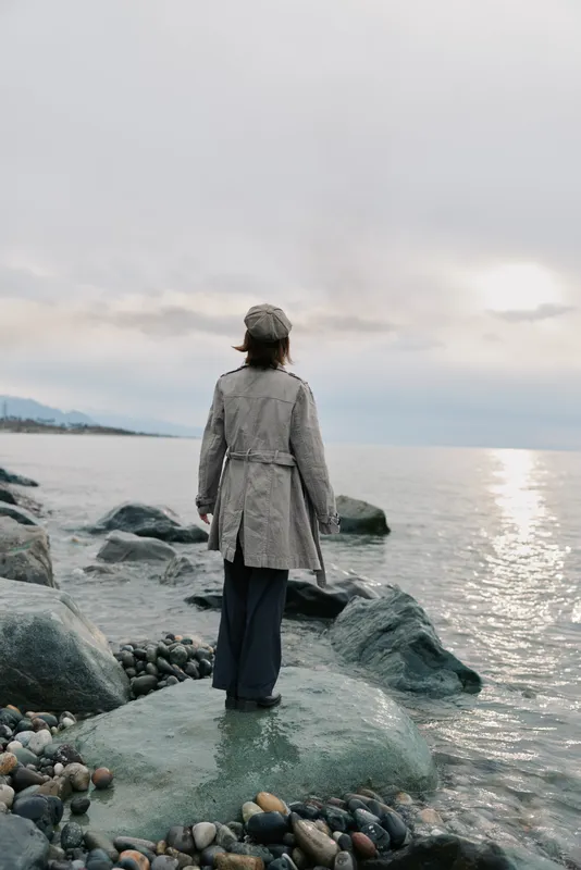 A woman wearing a coat and hat stands on a rocky shore looking at the calm sea under a cloudy sky. The scene conveys solitude, contemplation, and peaceful nature.