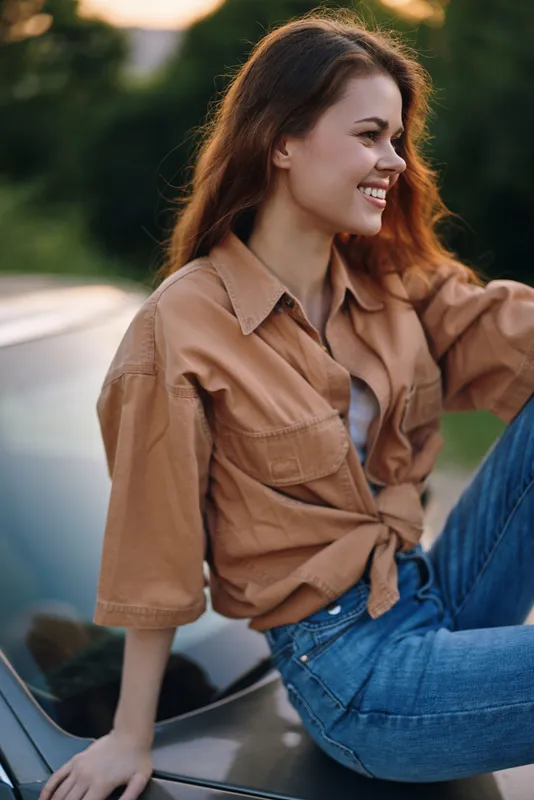 Smiling young woman in casual outfit sitting on a car, enjoying a sunny day outdoors with a warm background, embodying happiness and freedom in summer.
