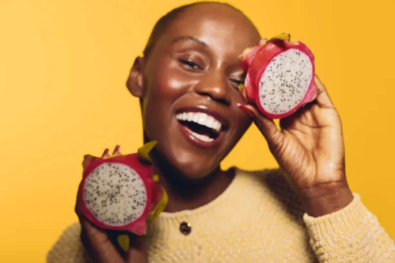 Happy dark skinned African woman holds two halves of dragon fruit near face smiling with teeth on yellow background. Concept of healthy lifestyle, exotic fruit, joy and nutrition.