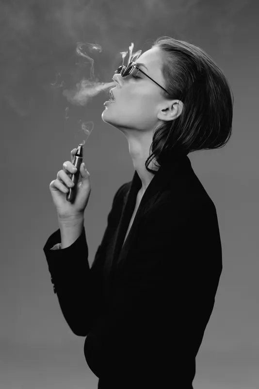 Black and white portrait of a stylish woman vaping with smoke in a minimalistic studio setting.