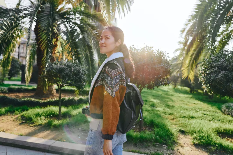 Woman with backpack strolls through a sunny park, embracing outdoor vibes, nature colors and casual fashion under palm trees and soft light, capturing a relaxed lifestyle moment