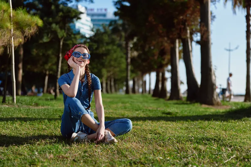 lifestyle woman in modern boho-western denim street style sitting on grass with sunglasses and braids, film color mood sunny day outdoors with green trees background