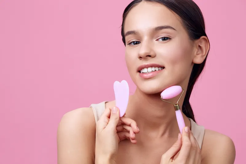 Beautiful young woman using facial tools on a pink background, showcasing skincare routine and promoting healthy skin care practices