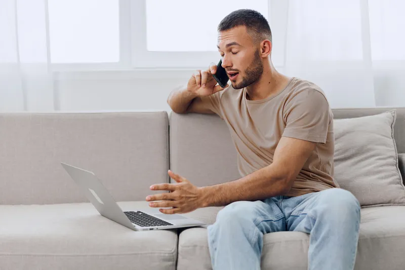 Productive man talking on the phone while working on a laptop at home. This modern lifestyle representation highlights remote work and effective communication.