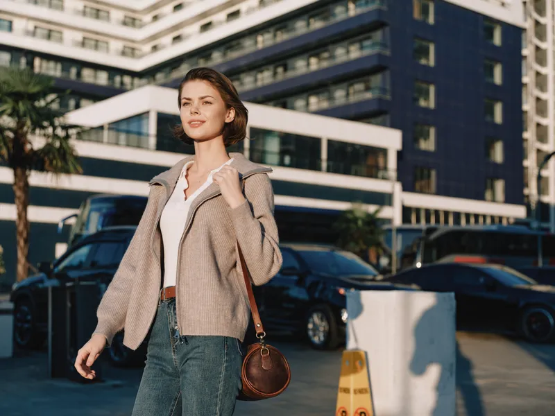 A cheerful professional woman walks along a sunny city street beside a modern office building, carrying a brown crossbody bag, wearing a light jacket and white shirt, exuding confidence.