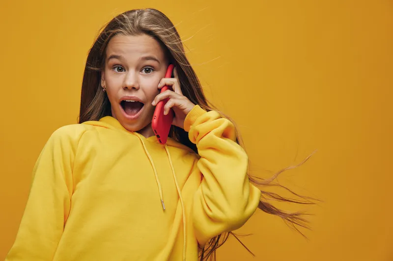 Girl with long hair wearing yellow hoodie talking on smartphone with surprised face expression on yellow background for communication and technology concept.