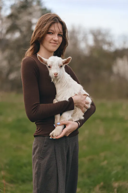 Young woman with long brown hair holds a white baby goat in her arms outdoors with a gentle smile during a casual lifestyle moment in nature.