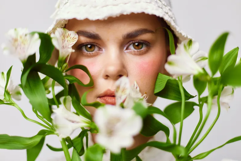 Young woman with a flower bouquet, wearing a white hat and natural makeup, conveying serenity and beauty with soft colors and a light background