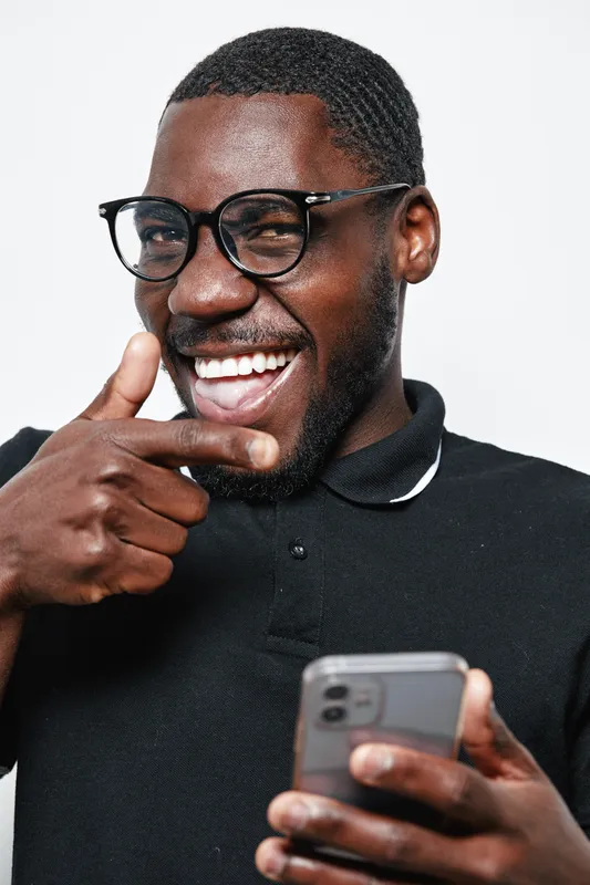 Black man with dark skin wearing glasses and black polo shirt smiling and pointing finger while holding smartphone in hand on white background. Positive expression and casual style.