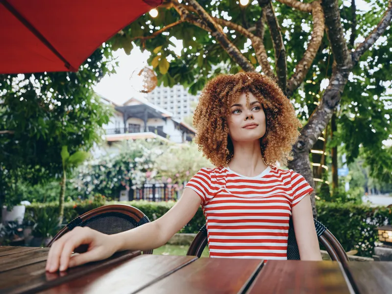 Portrait of a woman with curly hair seated at an outdoor cafe, wearing a red striped shirt, leaning on a wooden table under a red umbrella in a sunny garden setting.