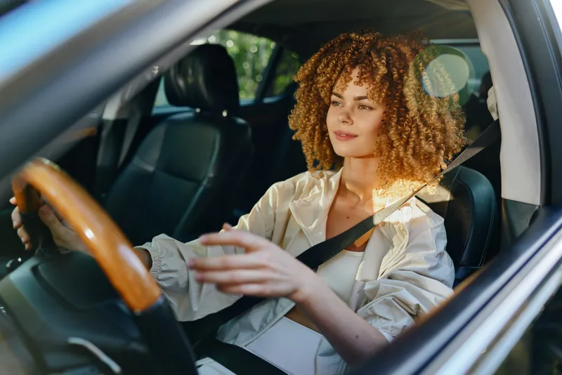 Woman with curly hair driving a car wearing a seatbelt, enjoying a sunny day and natural light, relaxed and focused on the road ahead.