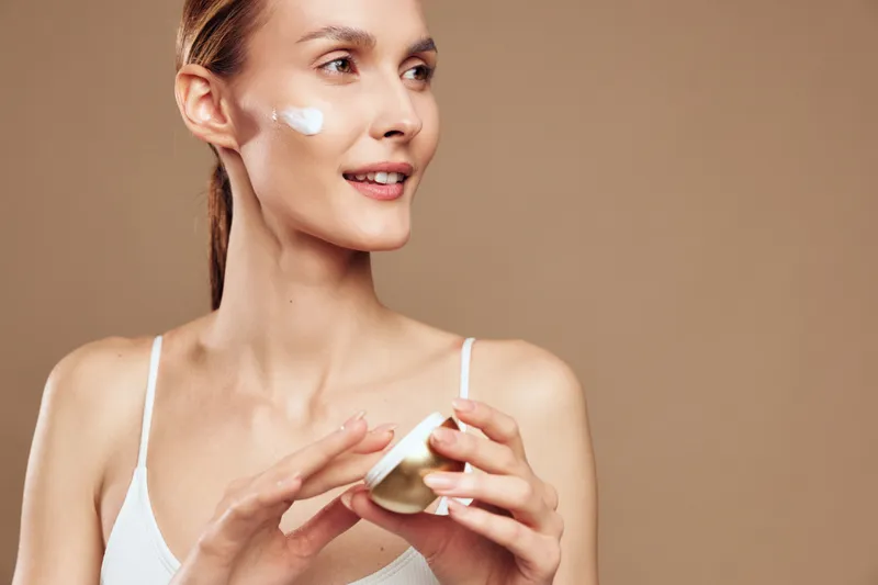 Young woman with clear skin applies moisturizing cream on her face holding an open jar smiling gently isolated on beige background skincare beauty concept.