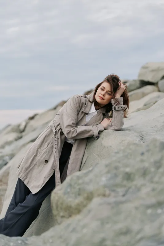 Young woman in a long coat leans on large rocks outdoors under cloudy sky. Natural light highlights casual fashion and thoughtful mood in a serene coastal environment.