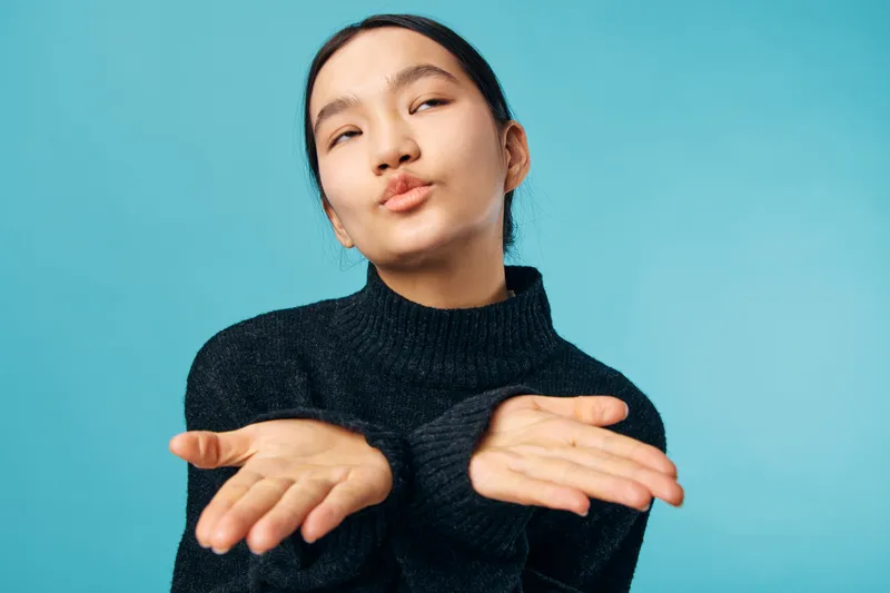 Young woman with black sweater blowing kiss with both hands, facial expression playful with eyes half closed on blue background in studio portrait closeup.