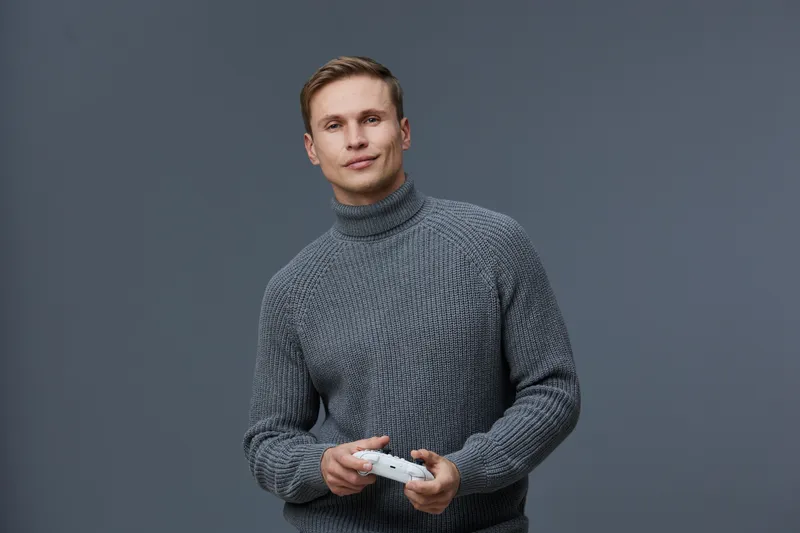 Man holding remote control in hand, looking at camera, smiling, casual clothing, grey background, studio portrait, positive facial expression, relaxed pose, modern style, lifestyle concept