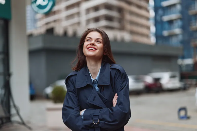 Young woman in a navy trench coat smiles happily while standing on a city street with her arms crossed, looking up with a confident and joyful expression.