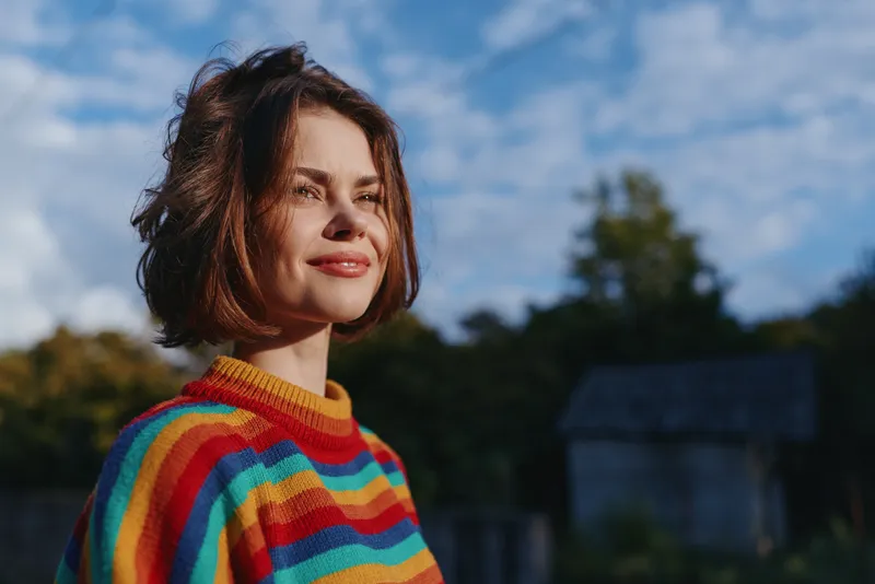 Woman smiling in a colorful striped sweater, short brown hair, young adult portrait outdoors in sunlight; relaxed gaze and casual outfit in rural countryside with travel lifestyle vibe.