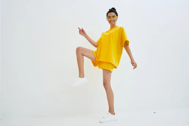 smiling woman in bright yellow casual outfit poses energetically against white background, wearing matching shorts and sneakers with cheerful expression