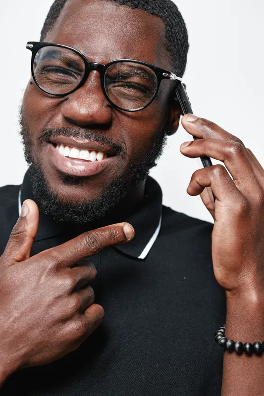 African American man with dark skin wearing glasses and black polo shirt smiling and talking on smartphone with hand gesture isolated on white background closeup portrait.