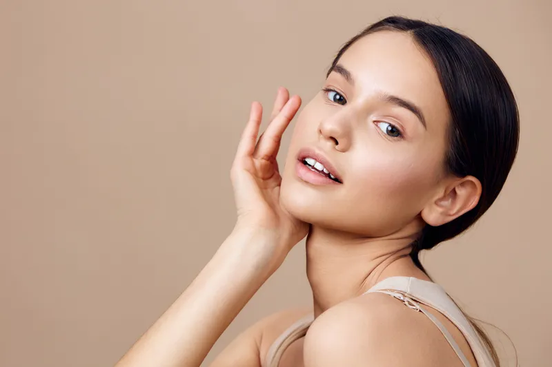Young woman with natural makeup and glowing skin smiling gently against a soft beige background, embodying beauty, confidence, and serenity