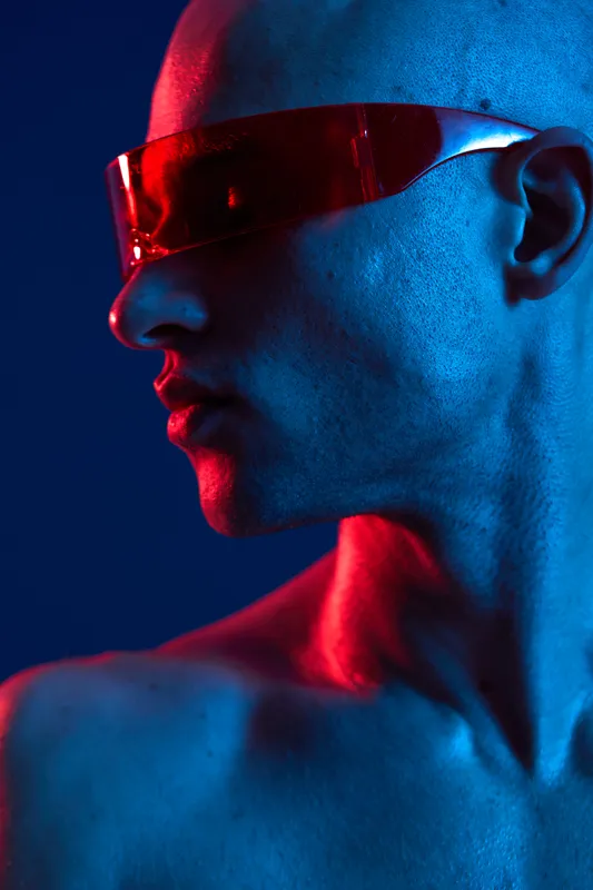 Fashion-forward man wearing futuristic sunglasses, showcasing a striking blue and red lighting contrast. Captivating expression enhances the modern vibe.