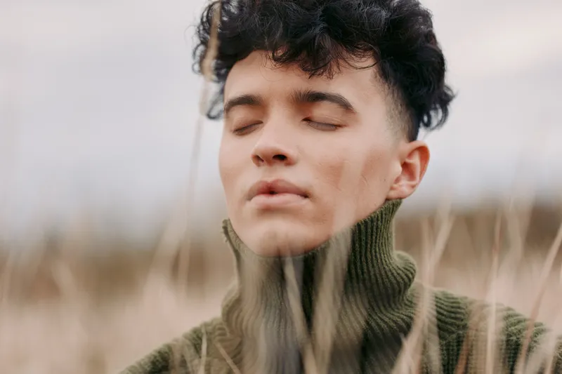 Serene portrait of a young person with eyes closed, wearing a knit turtleneck, standing in a natural field; authenticity and credibility shine through calm presence.