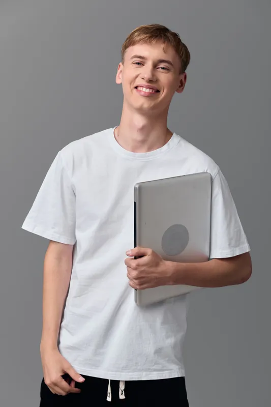 young man, student, laptop, casual, education, technology, smiling while holding a laptop and notebook in a studio for learning and career preparation in a modern creative setting