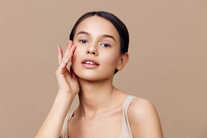 Portrait of a young woman with natural makeup and a serene expression against a warm beige background, emphasizing beauty and skincare
