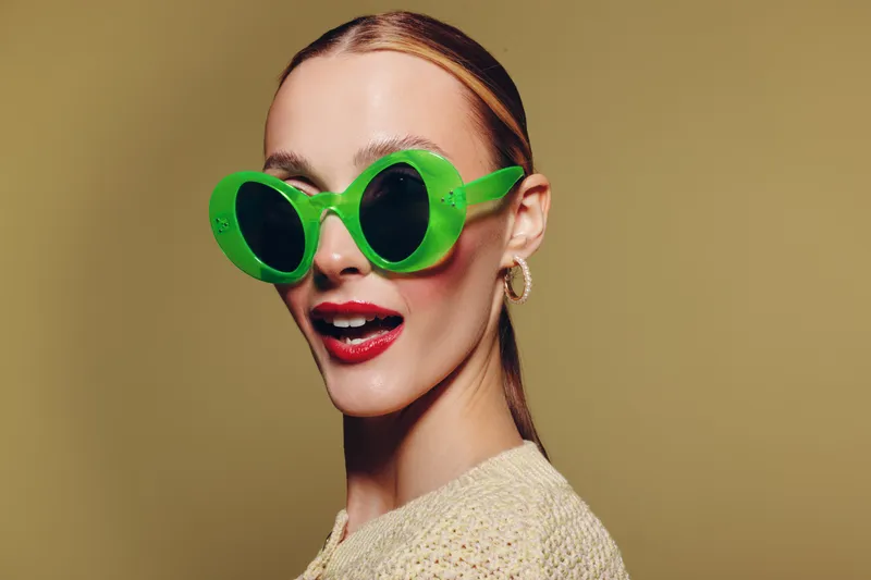 Stylish woman with slicked hair wearing large green sunglasses and red lipstick poses against beige background. Fashion portrait with bold accessories and confident expression.