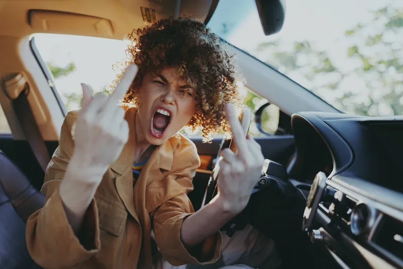Young woman with curly hair sits in a car and shouts with a bold rebellious mood, flipping the middle finger toward the camera during a spontaneous edgy street moment.