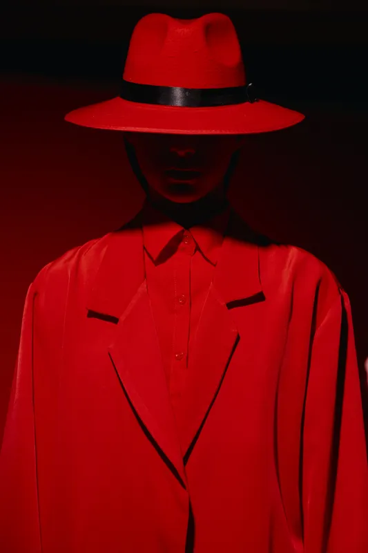 Mysterious person dressed in bold red outfit including wide-brimmed hat and oversized blazer under dramatic lighting. Concept for creative fashion editorial and beauty innovation.