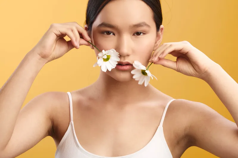 Asian woman gazes softly at the camera, holding white daisies near her cheeks against a warm yellow backdrop, a clean closeup portrait that conveys calm, freshness, and simple beauty.