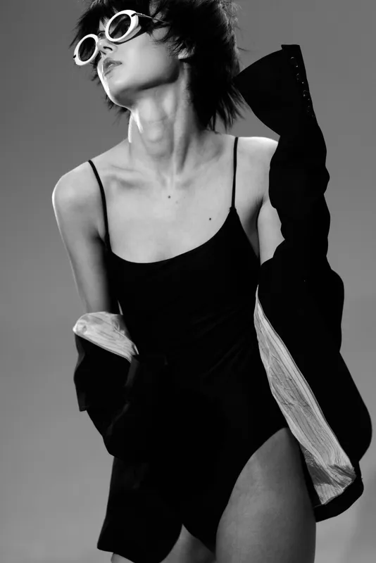 Black and white portrait of a woman wearing sunglasses and a black outfit in a stylish pose with dramatic lighting.