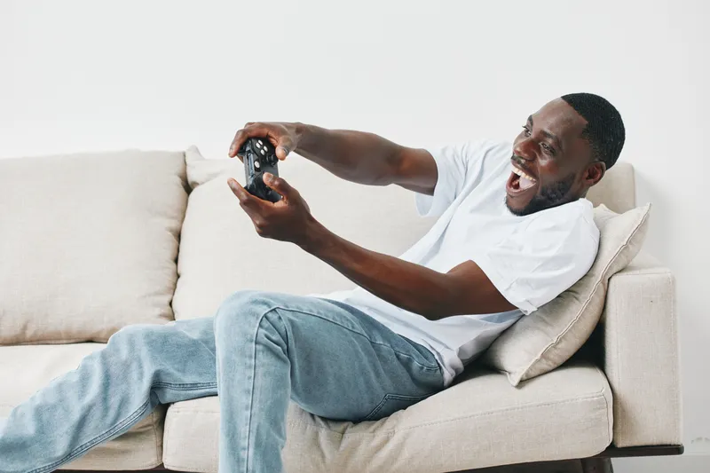Excited young Black man playing video games on a sofa, showcasing joy and engagement in a modern living room setting