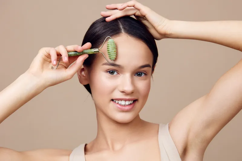 Young woman using a jade roller for facial massage, promoting healthy skincare routine with a natural look and soft smile, set against a neutral beige background