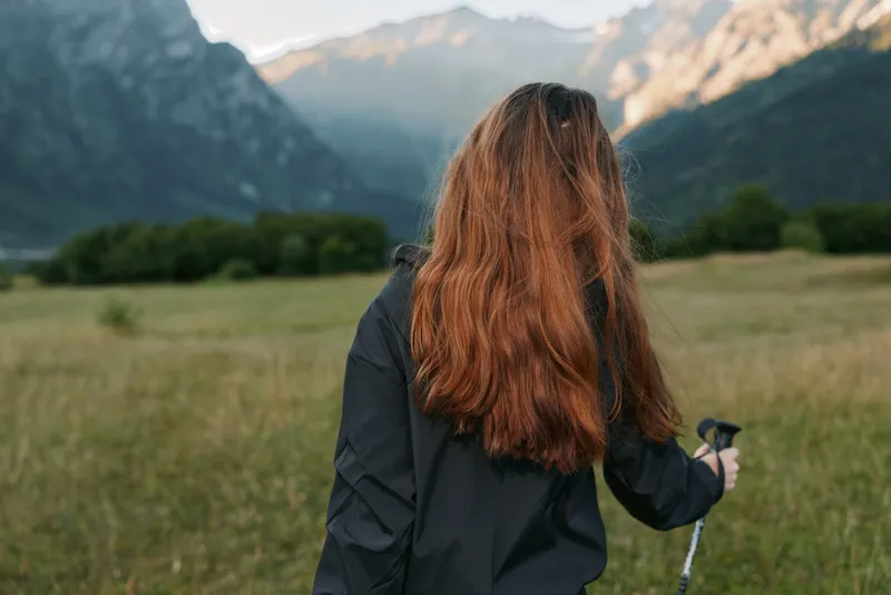 A lone hiker stands in a grassy meadow, facing distant mountains, holding a trekking pole. The landscape exudes adventure, calm, and open air.